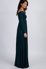 woman wearing a dark green off-shoulder maxi dress with long sleeves, standing sideways in a relaxed pose
