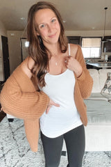 woman wearing a white tank top, black leggings, and an oversized brown knitted cardigan, smiling and posing indoors