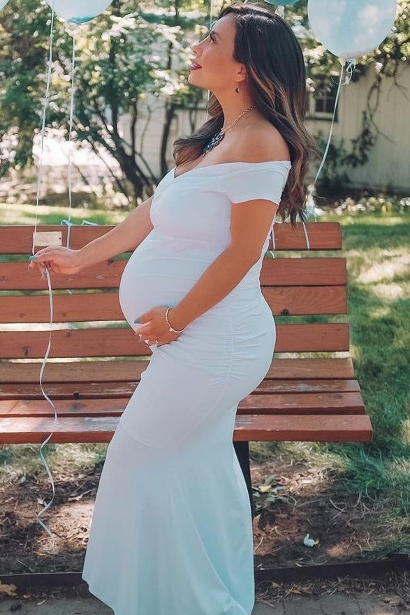 woman wearing a white off-shoulder maternity gown with a visible baby bump, standing outdoors by a bench
