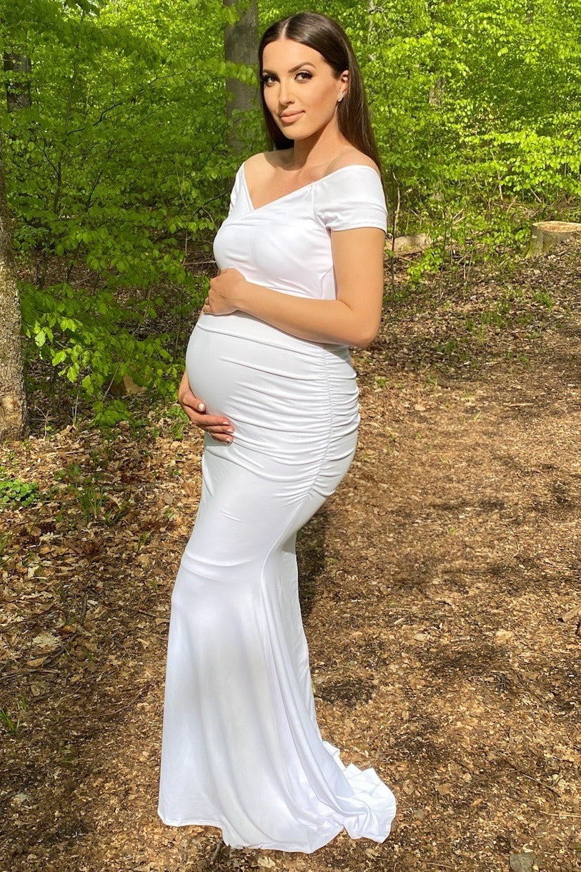 woman wearing a white off-the-shoulder maternity gown, showcasing her baby bump in a forest setting