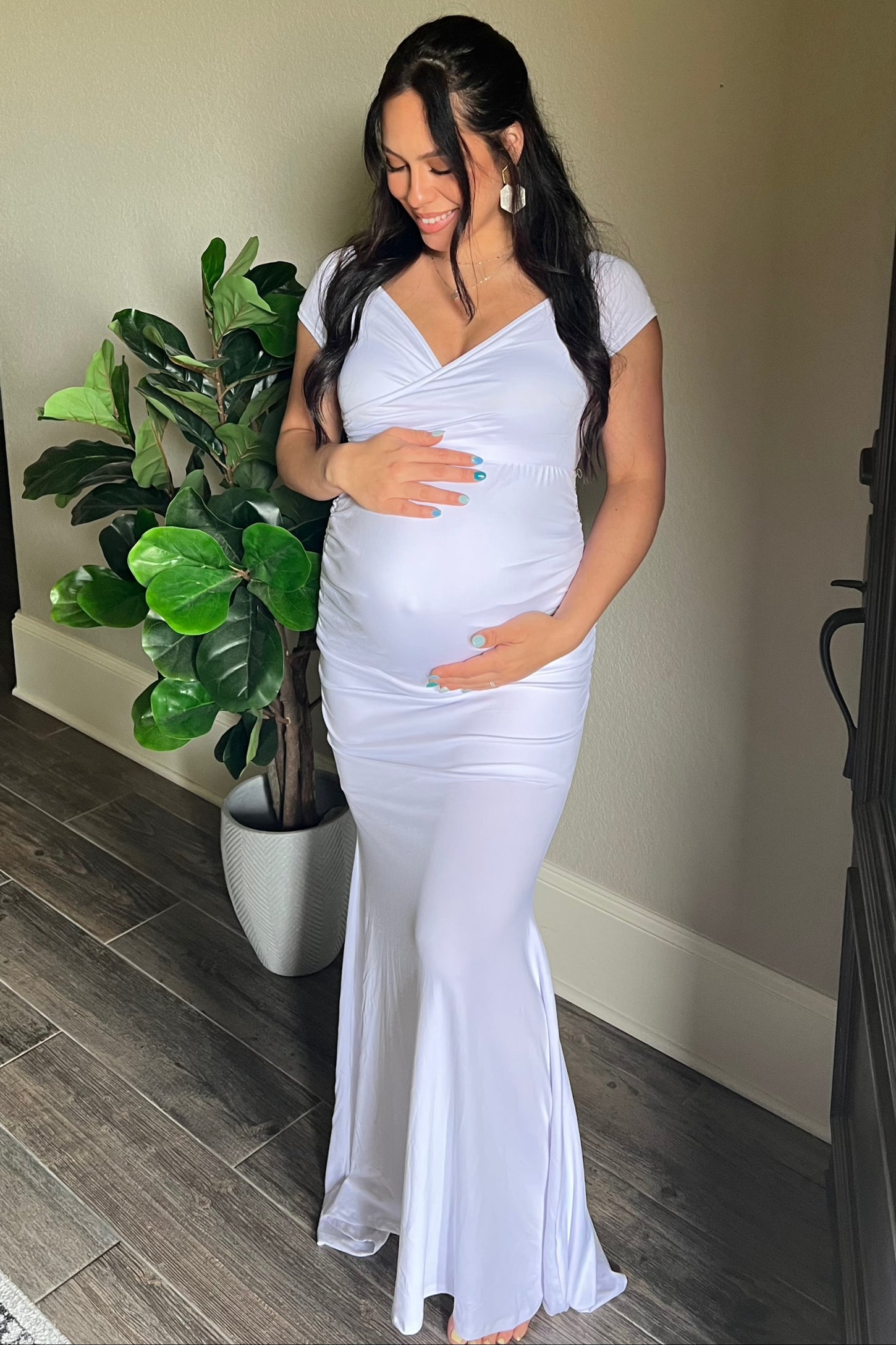 woman wearing a white maternity gown with a fitted design, holding her baby bump and standing beside a plant