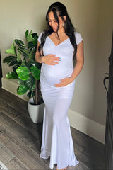 woman wearing a white maternity gown with a fitted design, holding her baby bump and standing beside a plant