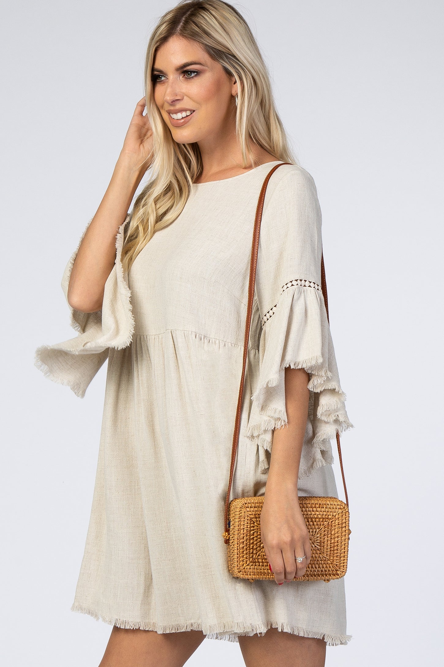 woman wearing a beige bohemian style dress with bell sleeves and fringe details, holding a woven crossbody bag