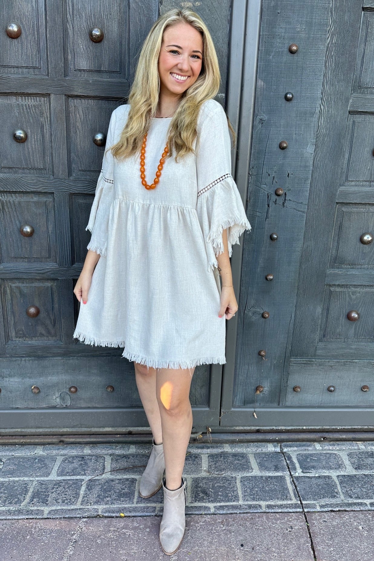 woman wearing a light gray bohemian-style dress with fringe details and bell sleeves, paired with an orange necklace and ankle boots