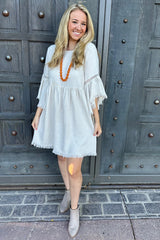 woman wearing a light gray bohemian-style dress with fringe details and bell sleeves, paired with an orange necklace and ankle boots