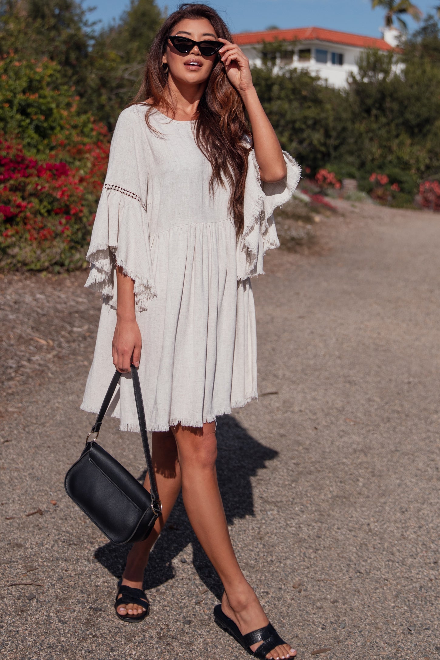 woman wearing a light beige short sleeve dress with flared sleeves and fringe details, carrying a black handbag