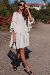 woman wearing a light beige short sleeve dress with flared sleeves and fringe details, carrying a black handbag