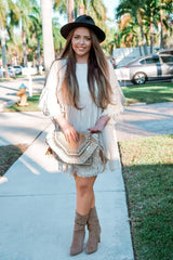 woman wearing a light beige fringe dress and high-heeled boots, holding a straw clutch, standing outdoors in a sunny setting