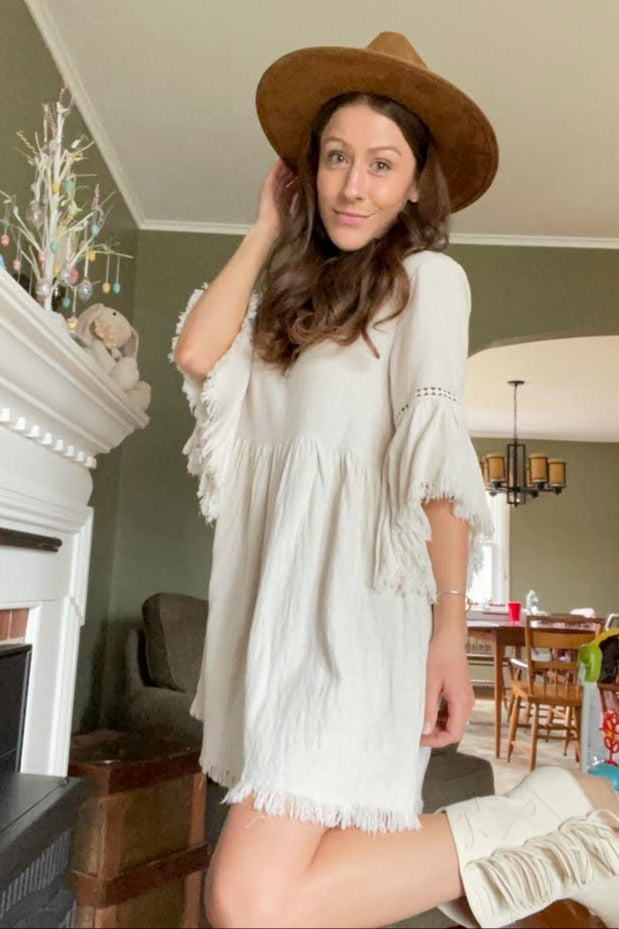 woman wearing a cream colored short sleeve dress with fringe detailing and a wide-brimmed hat