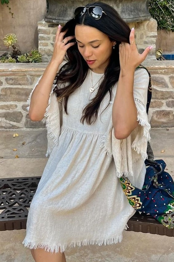 woman wearing a light beige short sleeve dress with fringe details and a relaxed fit, sitting outdoors