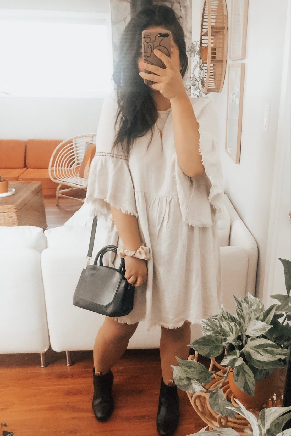 woman wearing a light beige loose fit dress with fringe sleeves and holding a black handbag in front of a mirror