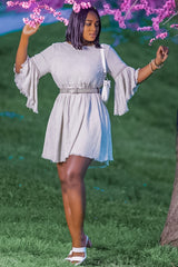 woman wearing a light gray belted dress with bell sleeves and frayed hem, standing in a garden with flowers