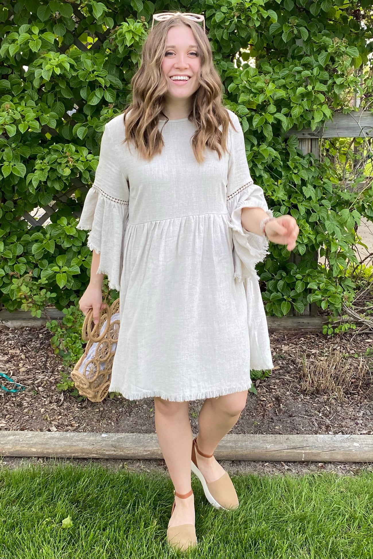 woman wearing a light beige tiered dress with ruffled sleeves and tassel details, smiling outdoors with greenery in the background