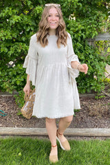 woman wearing a light beige tiered dress with ruffled sleeves and tassel details, smiling outdoors with greenery in the background