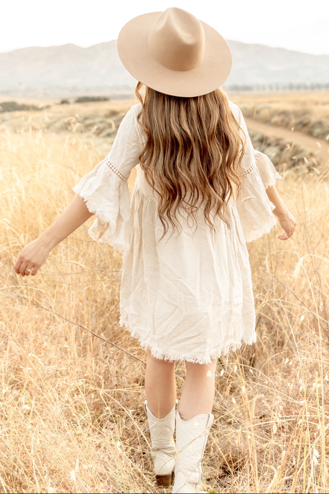 woman wearing a cream-colored bohemian-style dress with bell sleeves and fringed hem, walking through tall grass, wearing a wide-brimmed hat and cowboy boots