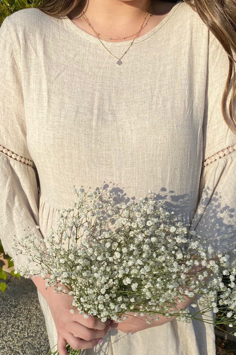 woman wearing a beige linen dress with bell sleeves, holding a bouquet of small white flowers
