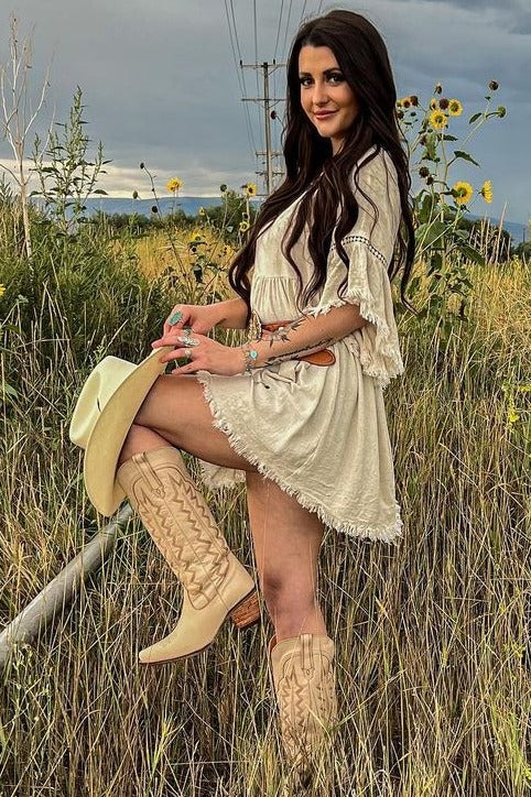 woman wearing a beige fringe dress and beige cowboy boots, standing in a field with sunflowers and holding a cowboy hat