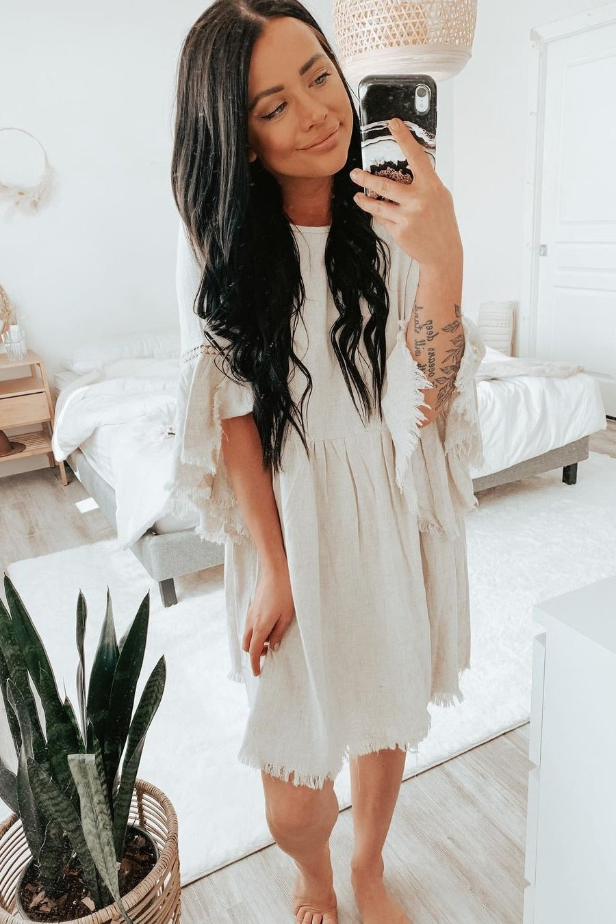 woman wearing a light beige short sleeve dress with frayed hem and bell sleeves, posing in a bright room with plants and a mirror