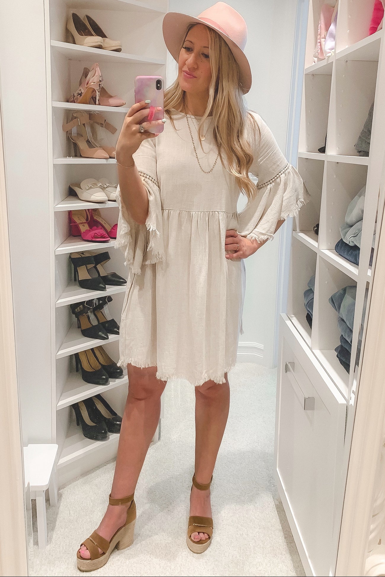 woman wearing a beige tiered dress with fringe sleeves and a pink hat, standing in a closet with shoes in the background