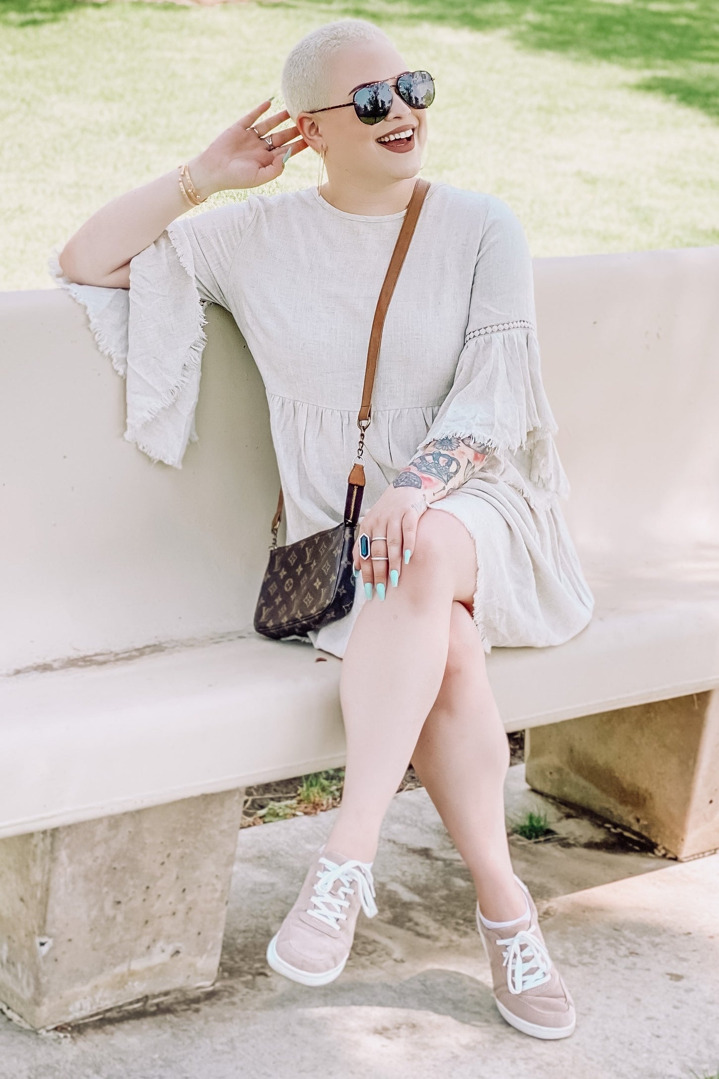 woman wearing a light beige flared sleeve dress and gray sneakers, sitting on a bench with sunglasses and a small handbag