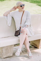 woman wearing a light beige flared sleeve dress and gray sneakers, sitting on a bench with sunglasses and a small handbag