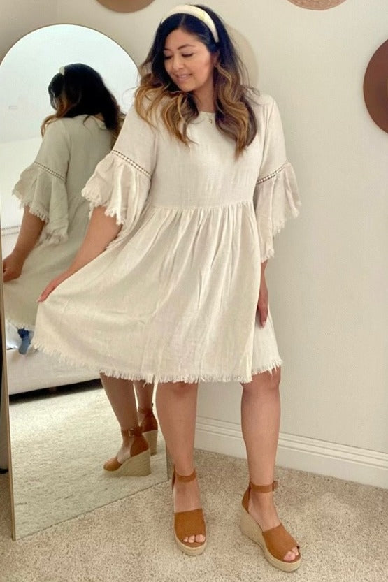 woman wearing a beige linen flutter sleeve dress and tan wedge sandals, posing in front of a mirror