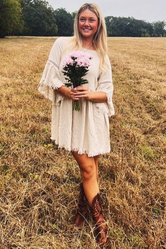 woman wearing a cream-colored boho dress with fringe and bell sleeves, holding a bouquet of flowers in a grassy field