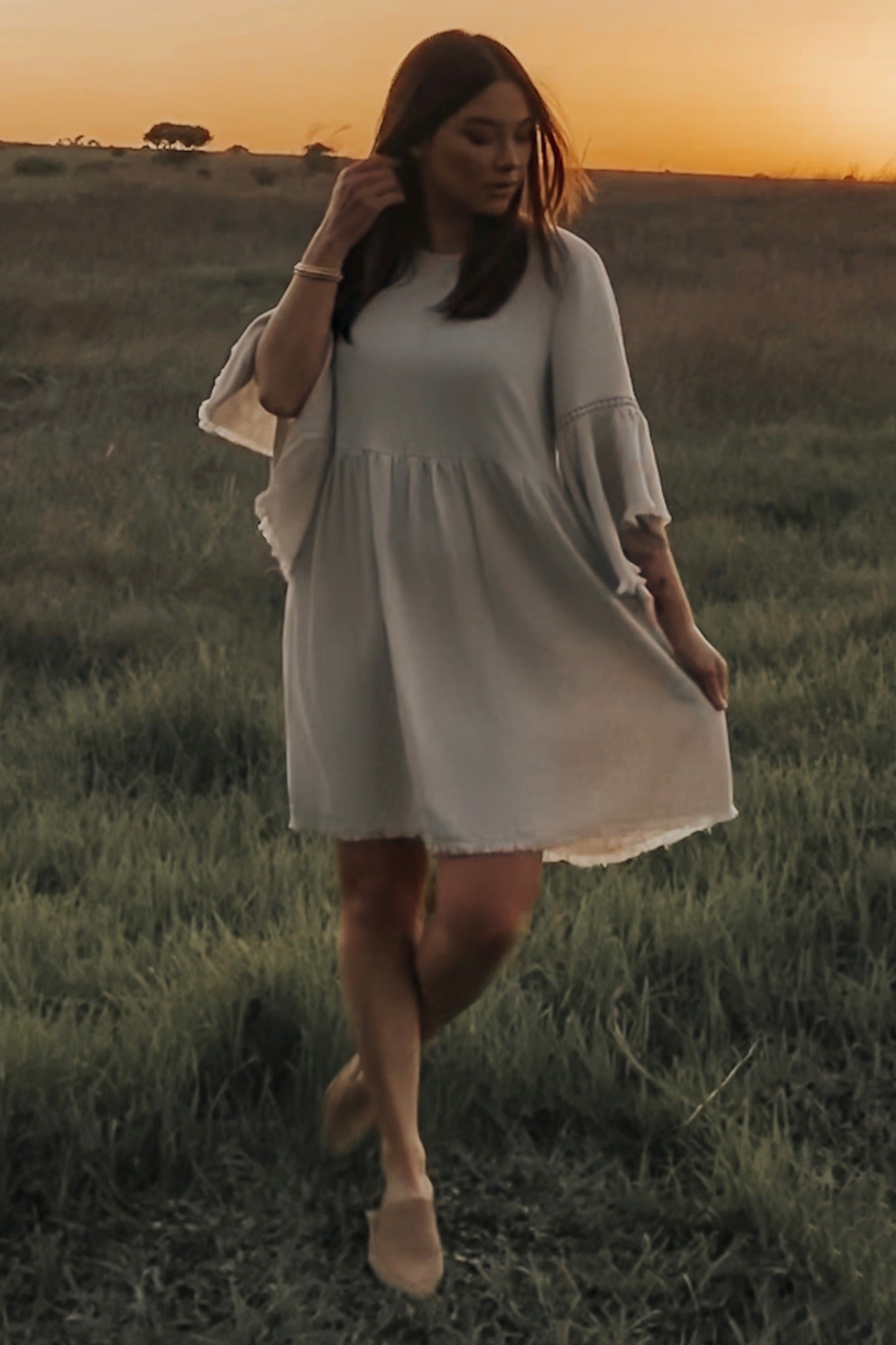 woman wearing a light gray flowy short sleeve dress standing in a grassy field at sunset