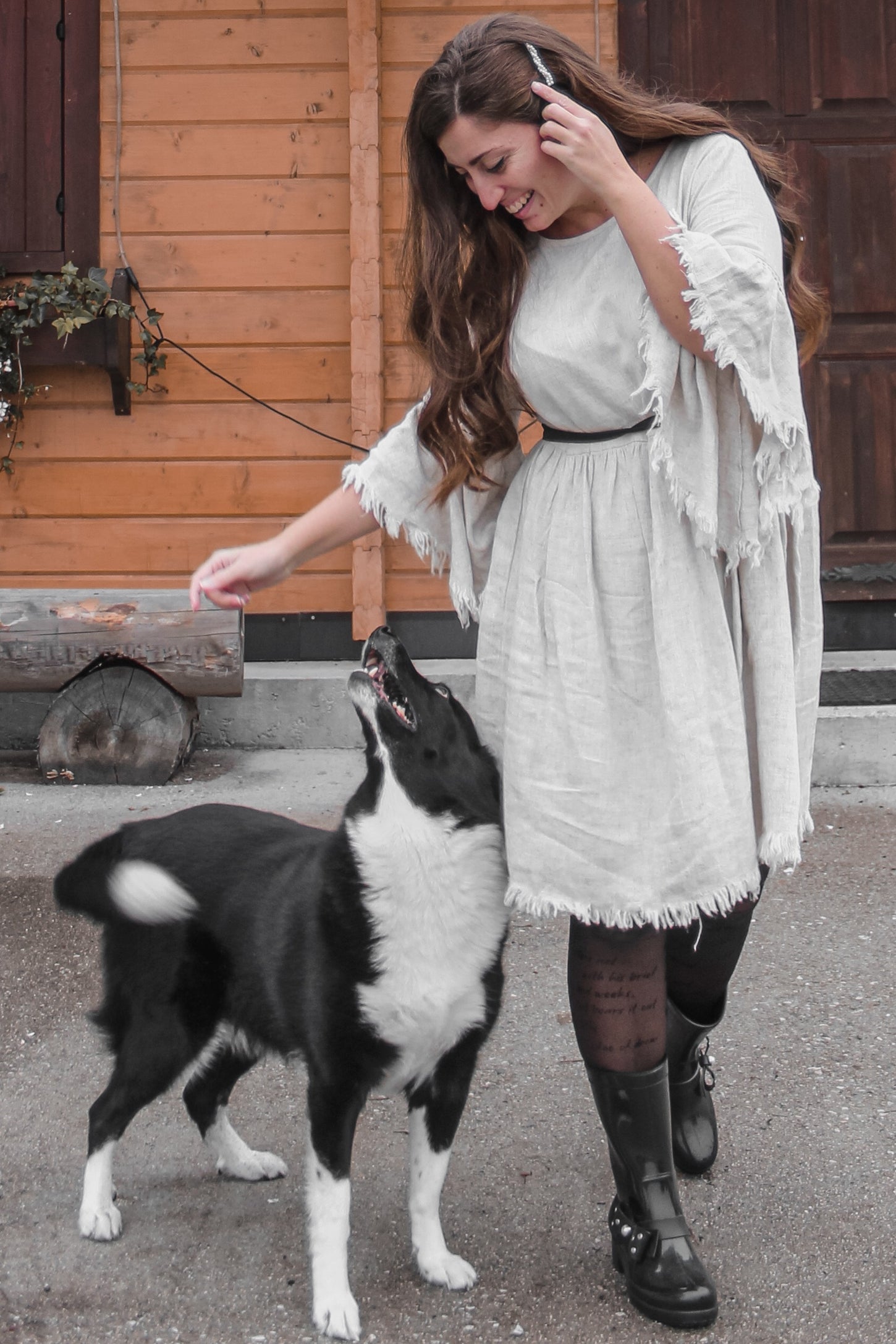 woman wearing a light gray fringed dress and black rain boots, petting a black and white dog outdoors