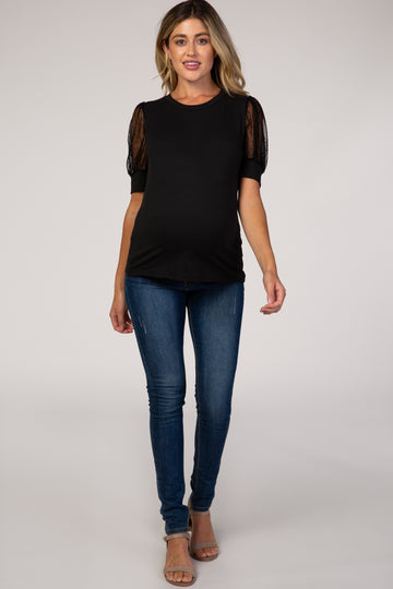 woman wearing a black maternity top with sheer puff sleeves and a visible baby bump, paired with blue skinny jeans