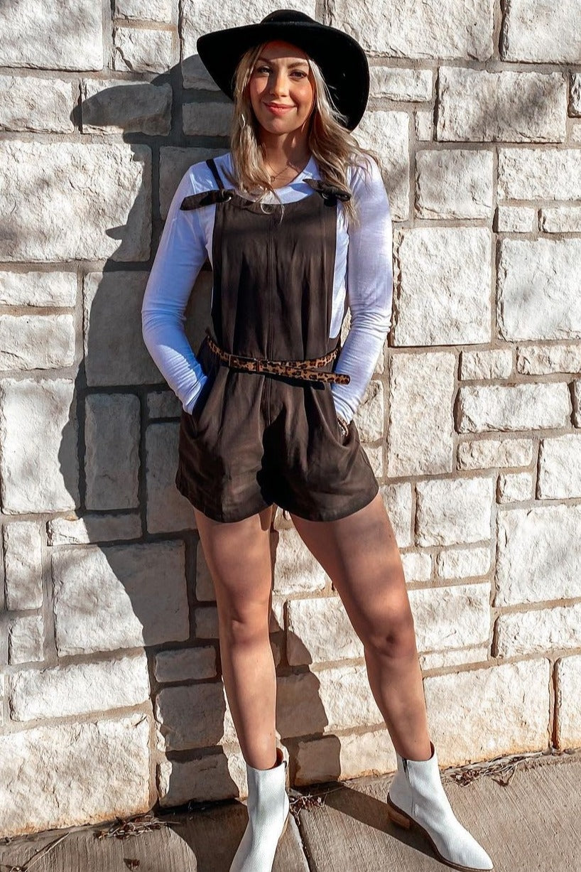 woman wearing a brown faux leather romper with a belted waist, styled with a white long sleeve top and a wide-brimmed hat