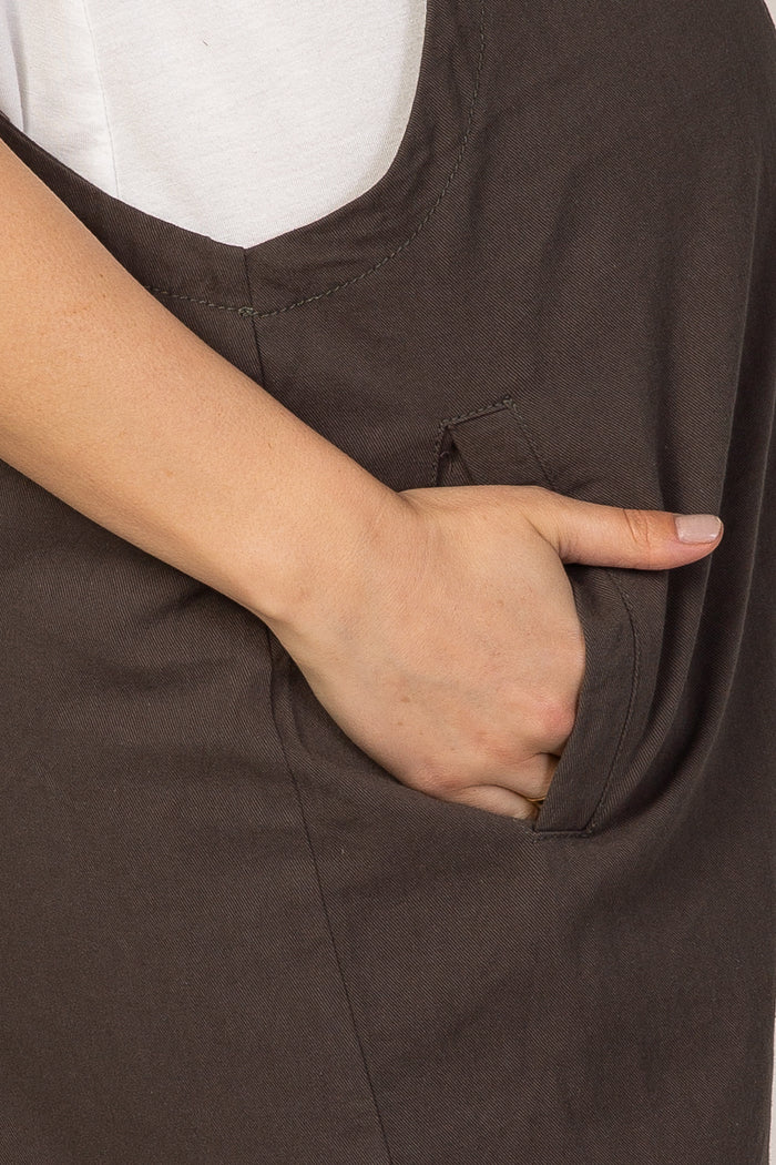 Close up of a hand in the side pocket of dark brown cotton overalls worn over a white t-shirt