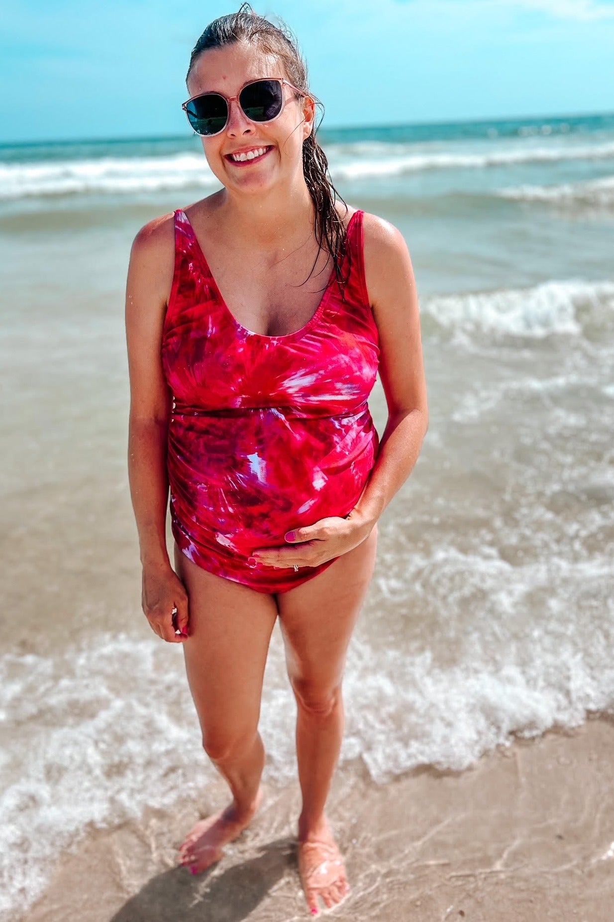 woman wearing a red tie-dye maternity swimsuit with a visible baby bump, standing in the ocean waves
