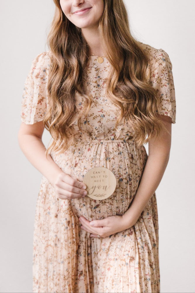 woman wearing a floral maternity dress holding a sign that says "can't wait to meet you" with her baby bump visible