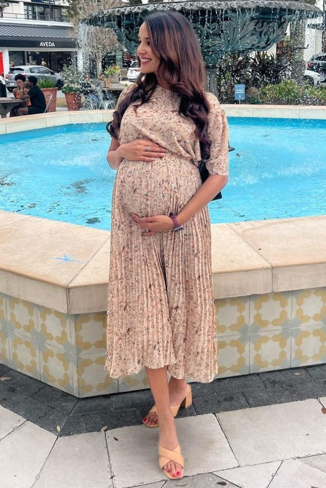 woman wearing a beige floral maternity dress with short sleeves and a visible baby bump, standing by a fountain
