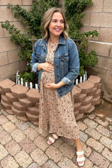 woman wearing a floral pleated maternity dress and denim jacket, holding her baby bump outdoors