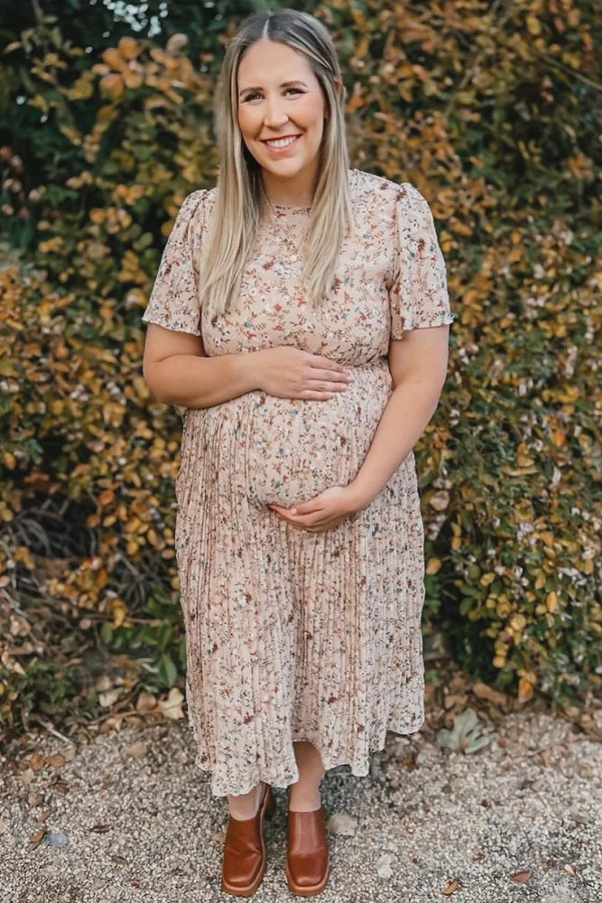 woman wearing a light pink floral maternity dress and holding her baby bump in an outdoor setting