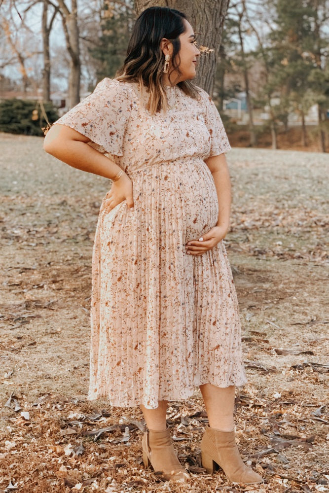 woman wearing a light pink floral maternity dress, holding her baby bump while standing outdoors