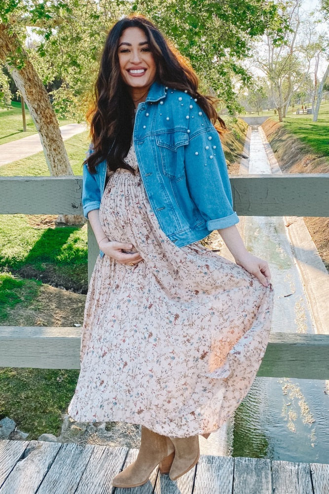 woman wearing a floral maxi maternity dress and denim jacket, holding her baby bump outdoors by a creek