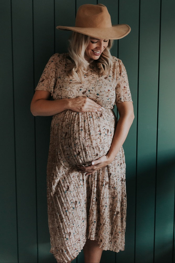 woman wearing a floral maternity dress and a wide-brimmed hat, gently cradling her baby bump