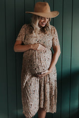 woman wearing a floral maternity dress and a wide-brimmed hat, gently cradling her baby bump