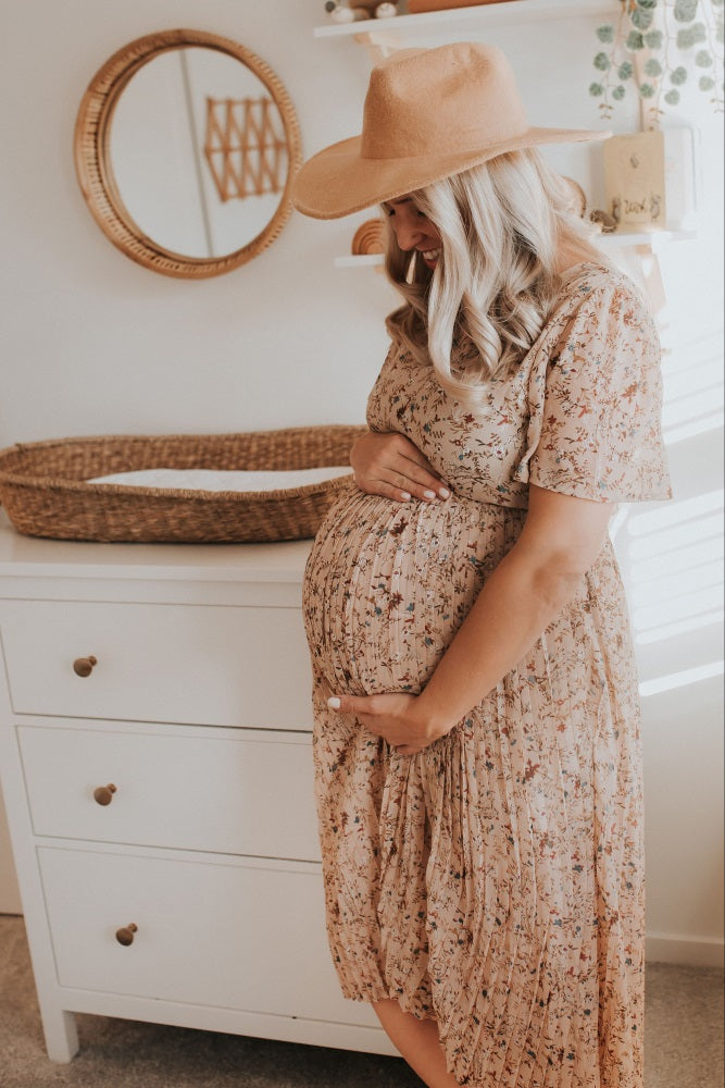 pregnant woman in floral dress and tan hat holding her belly in a nursery with white dresser and wicker basket