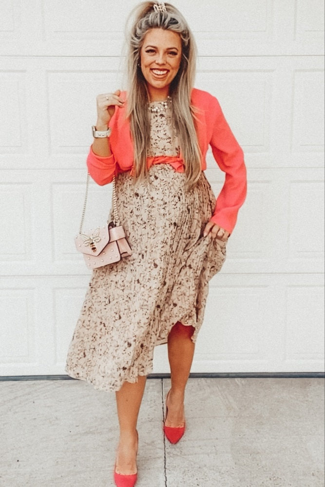 woman wearing a floral maternity dress with a coral blazer and holding a small pink handbag, smiling and showing baby bump