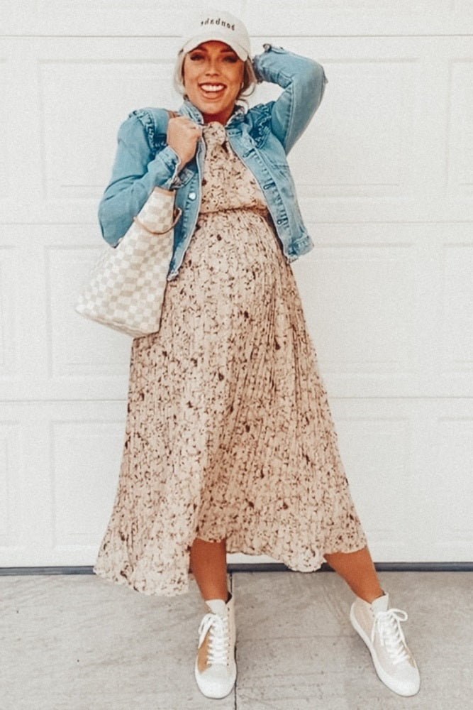 woman wearing a floral maternity maxi dress with a denim jacket and sneakers, holding a handbag and showcasing her baby bump