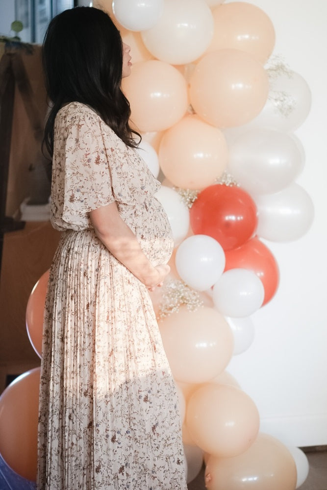 woman wearing a floral beige maternity dress and holding her baby bump with balloons in the background