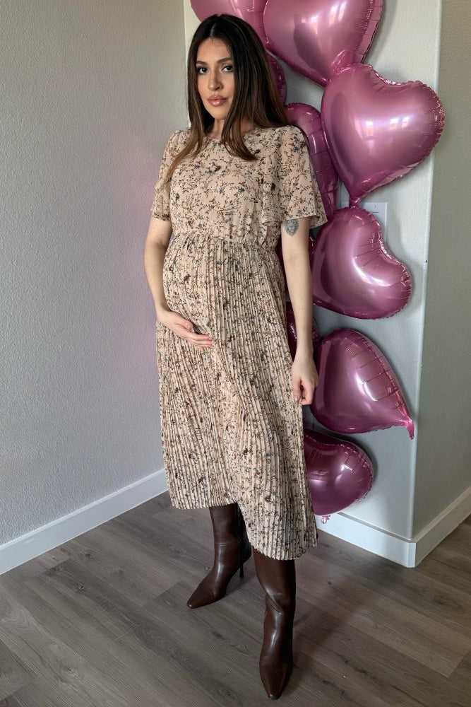 woman wearing a floral print maternity dress with pleats, holding her baby bump, standing in front of pink heart balloons