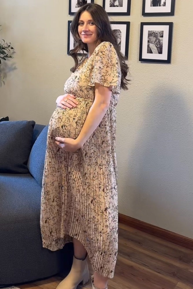 woman wearing a floral print maternity dress with a tiered skirt, holding her baby bump while standing by a sofa