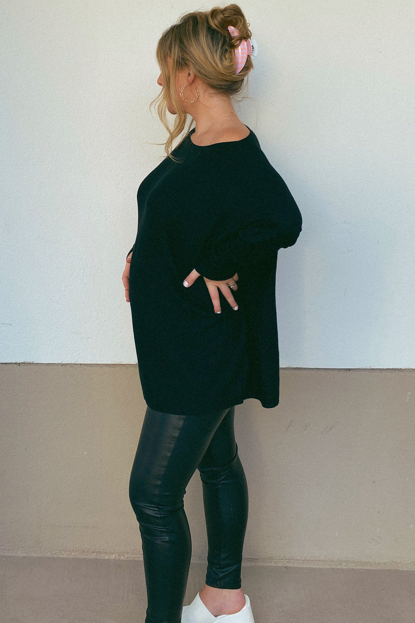 pregnant woman in black oversized sweater and faux leather leggings posing against a neutral wall