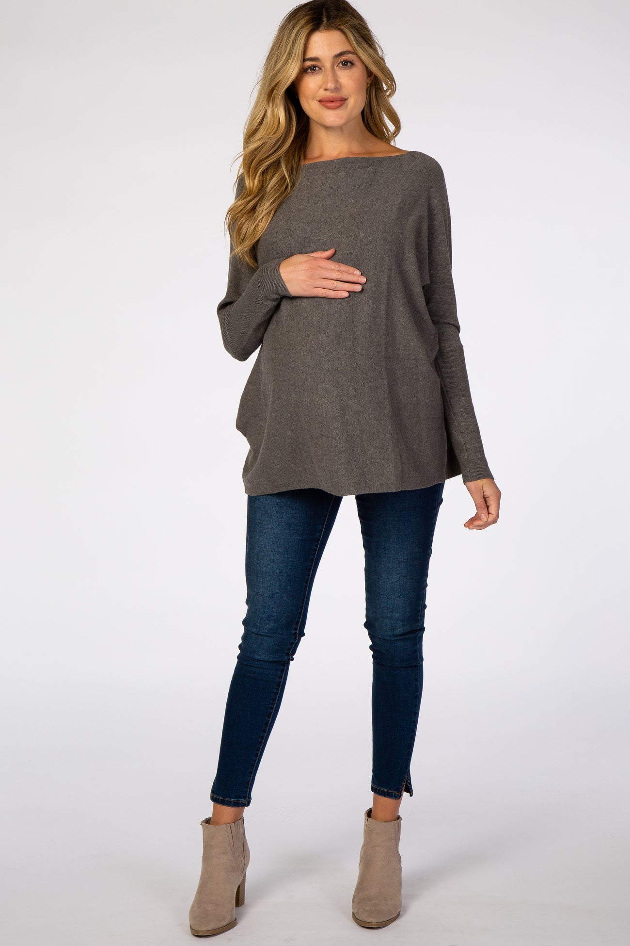 woman wearing a dark gray maternity sweater, holding her baby bump, paired with skinny jeans and ankle boots