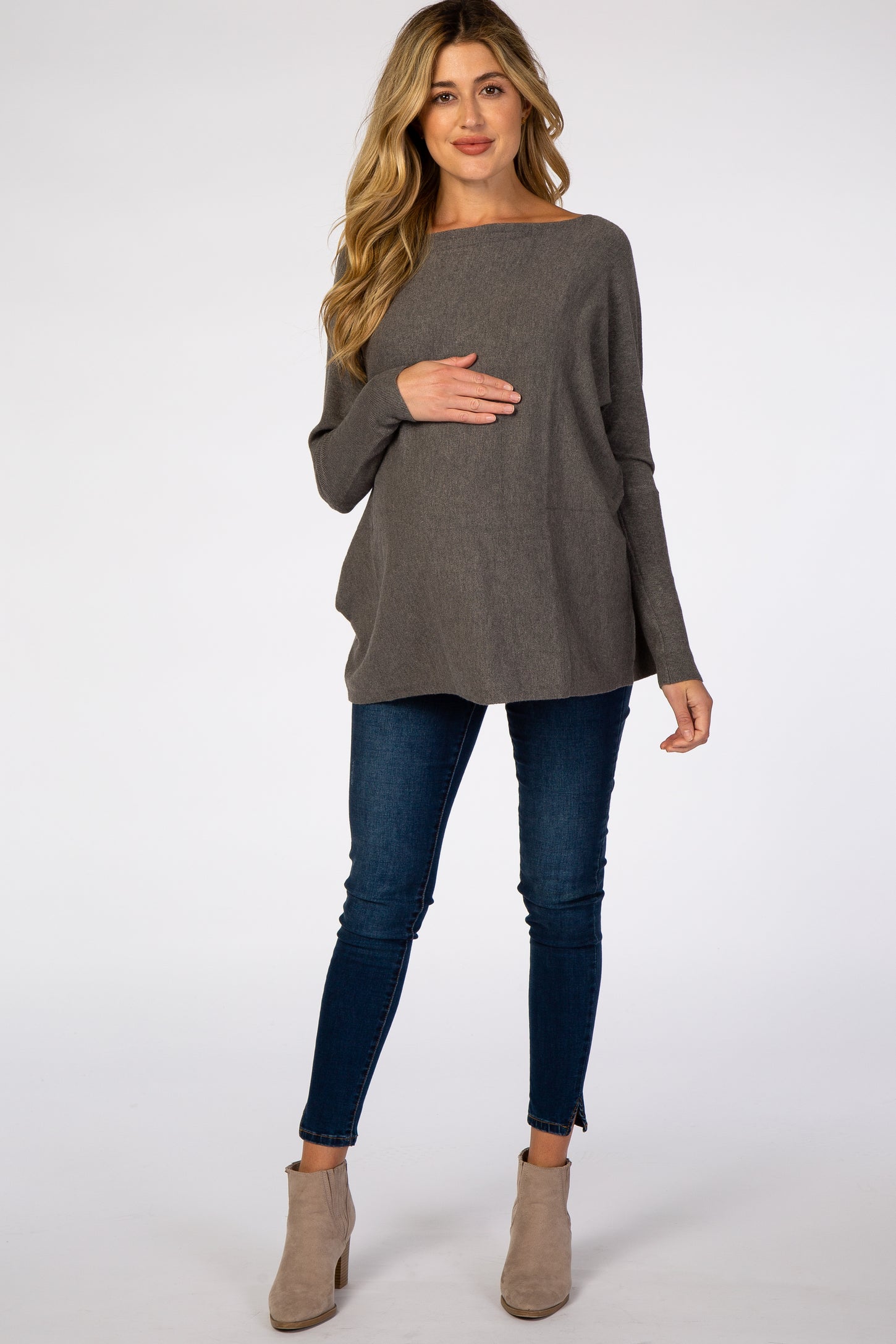 woman wearing a dark gray maternity sweater, holding her baby bump, paired with skinny jeans and ankle boots